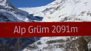 Ein rot-weißes Wegweiser-Schild vor einer imposanten, schneebedeckten alpinen Berglandschaft in der Schweiz.
