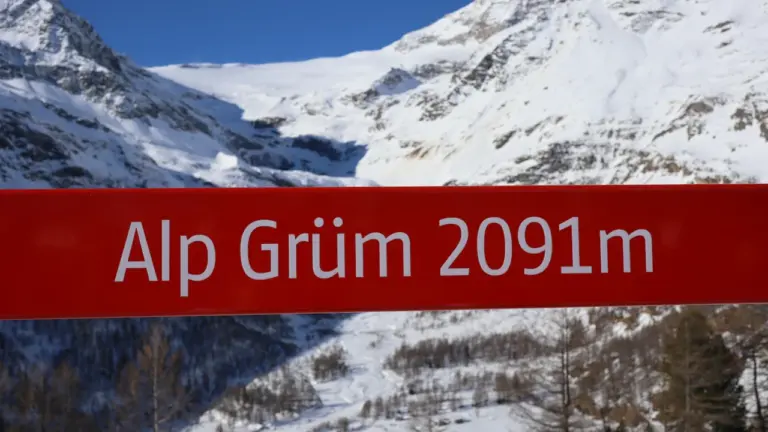Ein rot-weißes Wegweiser-Schild vor einer imposanten, schneebedeckten alpinen Berglandschaft in der Schweiz.