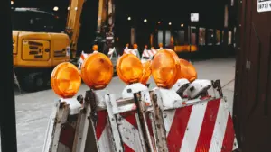 Orangefarbene Baustellenleuchten und rot-weiße Absperrungen vor einem Bagger in der Nacht.