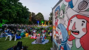 Zuschauer sitzen im Freien auf Decken vor einer beleuchteten Bühne in einem Park.