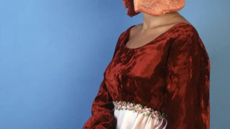 Eine Frau in einem historischen roten Kleid trägt ein Stück rohes Fleisch als Kopfbedeckung.