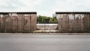 Ein zerfallener Mauerabschnitt in Berlin trennt eine leere Straße von grünem Gelände.