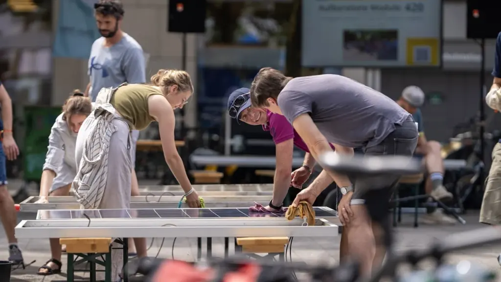 Mehrere Personen reinigen gemeinsam auf Bierbänken montierte Solarmodule in einem belebten städtischen Außenbereich.