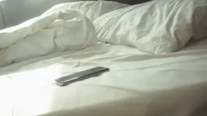 Ein einsames Smartphone liegt auf einem ungemachten Bett im hellen Schlafzimmer.