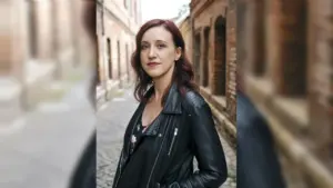 Eine junge Frau mit rötlichem Haar trägt eine schwarze Lederjacke in einer alten Backsteingasse.