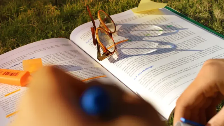 Ein aufgeschlagenes Lehrbuch mit Brille und Textmarkern liegt auf einer grünen Wiese.